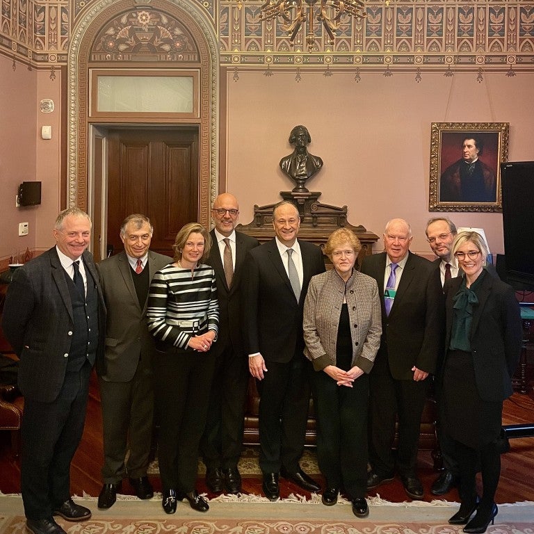 photo in the White House under a grand chandelier, pink background, row of people smiling at the camera: Lord John Mann, Katharina von schnurbein, ted Dutch, second gentleman Doug emhoff, Deborah Lipstadt, Andrew baker, Felix Klein, holly Huffnagle