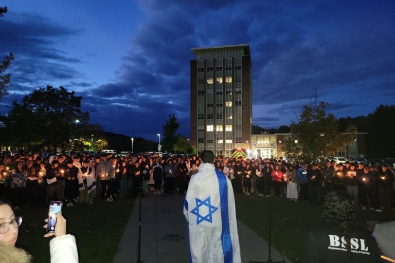 figure draped in an israeli flag, facing a crowd of students holding candles at a vigil at Binghamton University, for victims of October 7th Hamas terrorist attacks