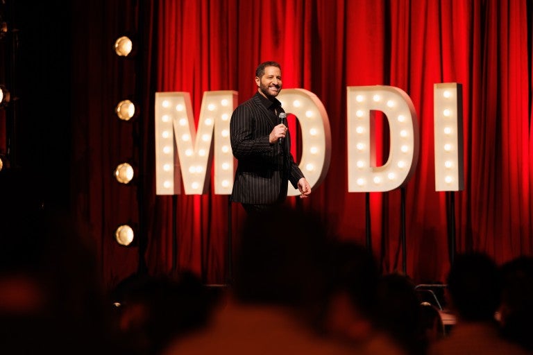 Red curtain background with marquee light up letters spelling MODI, Modi Rosenfeld walking across a stage, in a black suit, holding a microphone, smiling