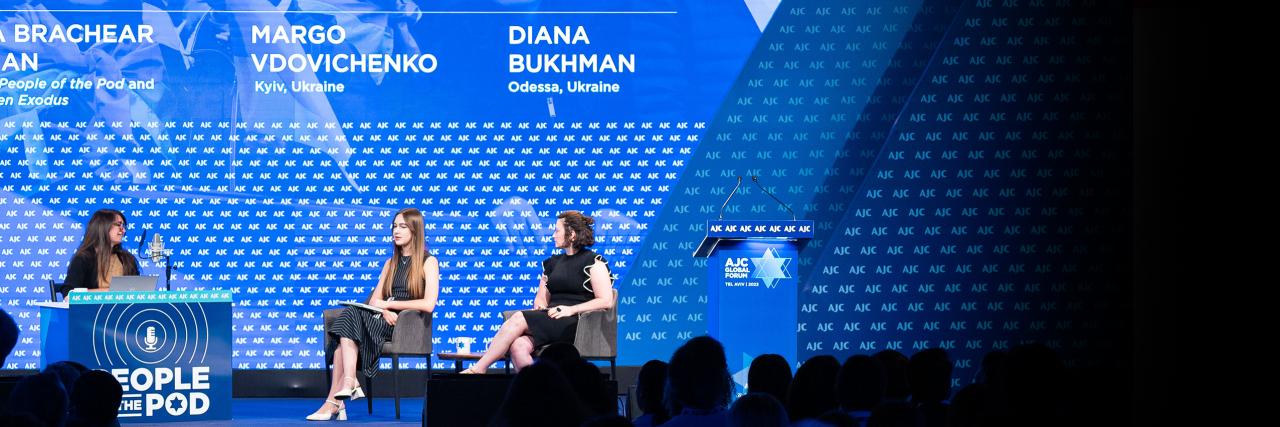 Three women on a stage with a blue background sitting in chairs, one behind a desk branded with People of the Pod