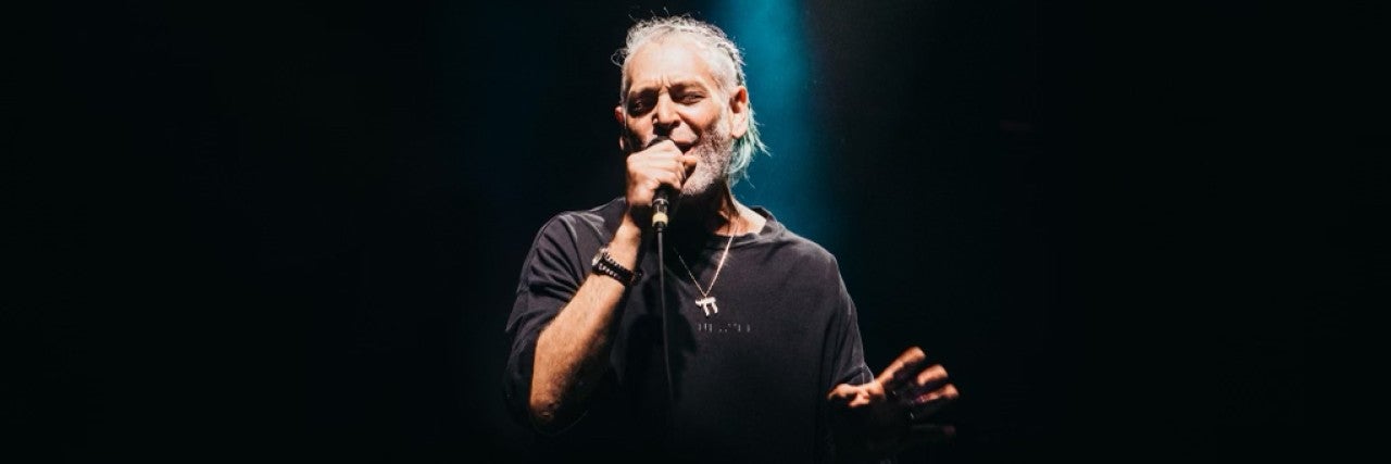 Singer Matisyahu on stage singing into a microphone, background is dark except for one spotlight directed down towards him. Matisuahu is wearing a dark shirt and a large silver necklace with a Chai pendant.