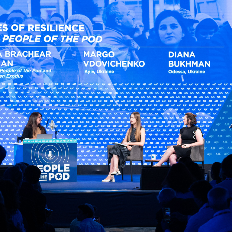 Three women on a stage with a blue background sitting in chairs, one behind a desk branded with People of the Pod