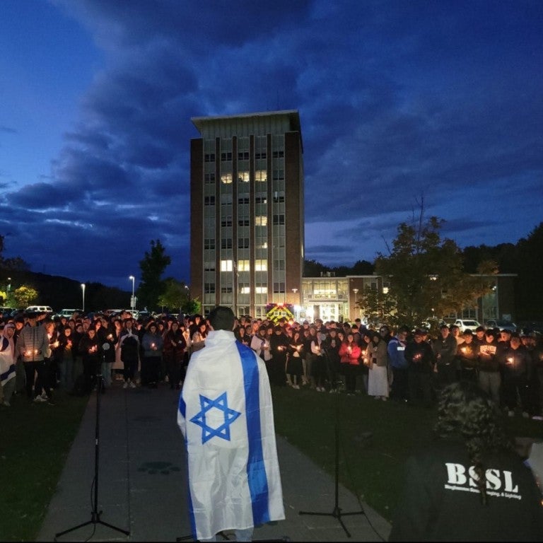 figure draped in an israeli flag, facing a crowd of students holding candles at a vigil at Binghamton University, for victims of October 7th Hamas terrorist attacks