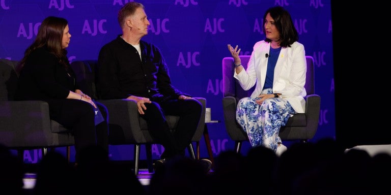 Three people sitting on chairs on a stage in front of a blue backdrop that reads AJC step and repeat. Belle Yoeli, Michael Rapaport, and Aviva Klompas