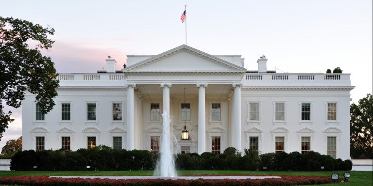 A scenic image of the White House with an American flag on the roof, featuring a fountain and green lawn.