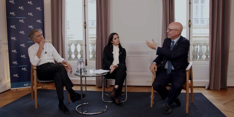 L-R: Bernard-Henri Lévy, Anne-Sophie Sebban-Bécache, and Ted Deutch, seated, in front of two windows and an AJC step and repeat background, in conversation