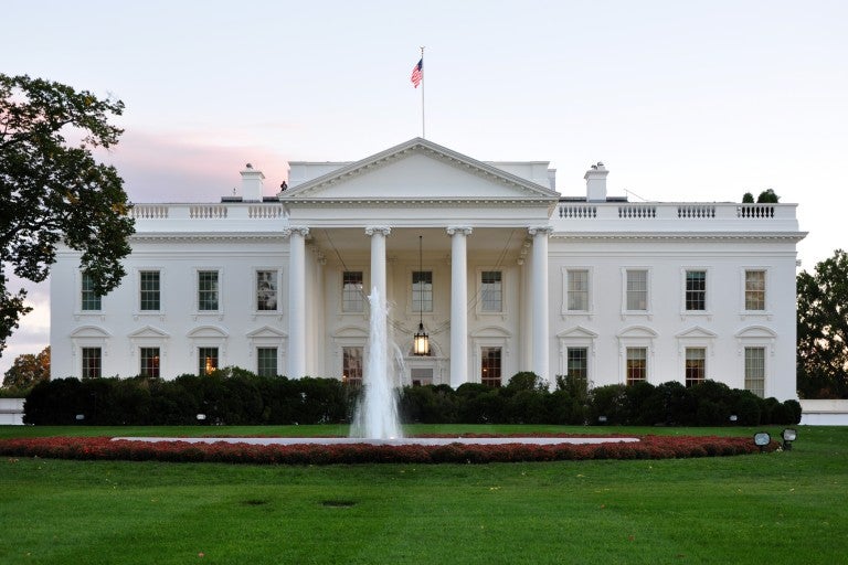 A scenic image of the White House with an American flag on the roof, featuring a fountain and green lawn.