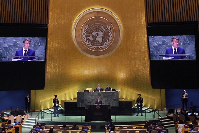 Large assembly hall filled with delegates features French President Emmanuel Macron speaking at a podium under the United Nations emblem, with two large screens displaying his image, during the UN General Assembly.