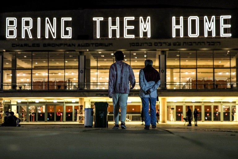 two figures standing in front of a building that is lit up with large white lettering spelling out 'bring them home' in all caps.