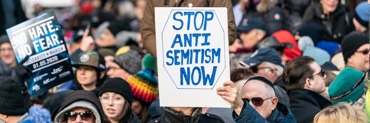 Photo of a crowd and a man holding a sign saying "Stop Antisemitism Now"