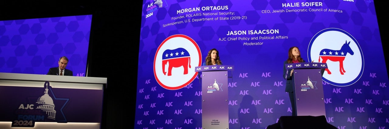 On a stage, there's a large desk and two podiums. A man, Jason Isaacson, is sat at the desk. Two women, Morgan Ortagus on the left and Halie Soifer on the right, stand at podiums on a stage with a backdrop that says "AJC Global Forum 2024" and "The Great Debate: Election 2020," followed by their names