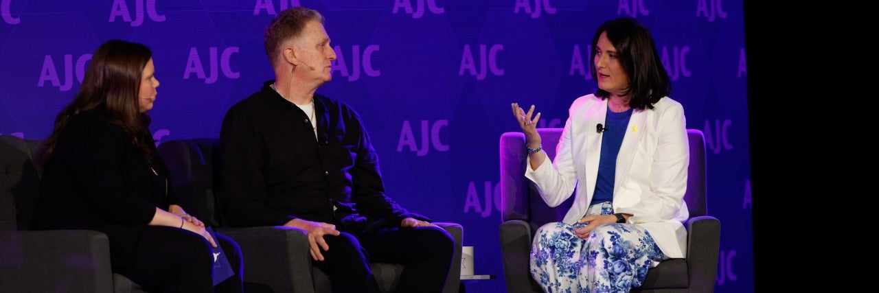 Three people sitting on chairs on a stage in front of a blue backdrop that reads AJC step and repeat. Belle Yoeli, Michael Rapaport, and Aviva Klompas