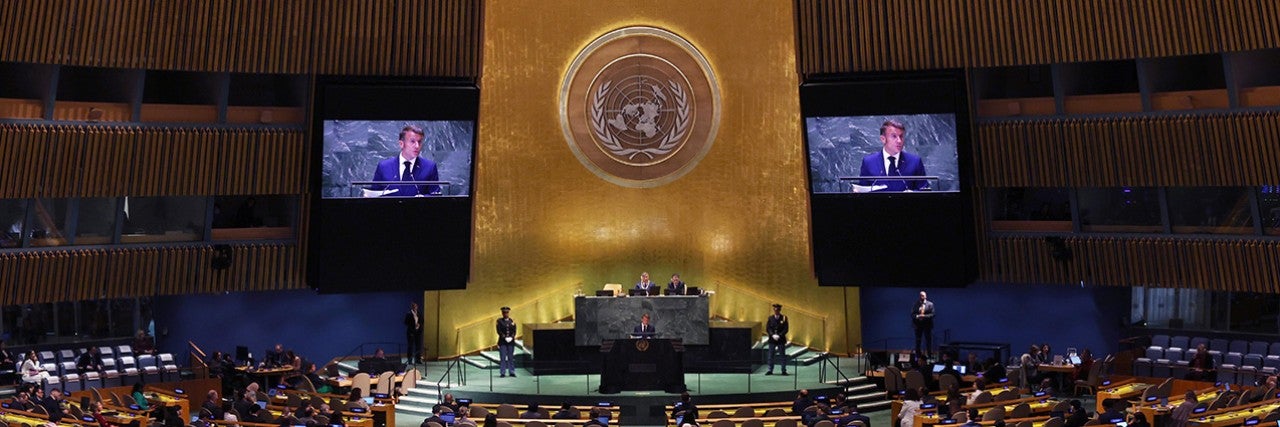 Large assembly hall filled with delegates features French President Emmanuel Macron speaking at a podium under the United Nations emblem, with two large screens displaying his image, during the UN General Assembly.