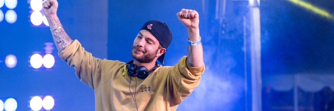 Israeli DJ Daniel Vaknin, in a yellow sweater stands with his arms raised, expressing joy or celebration. 