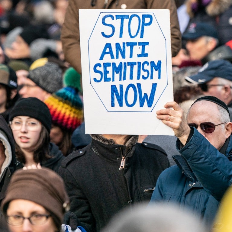 Photo of a crowd and a man holding a sign saying "Stop Antisemitism Now"
