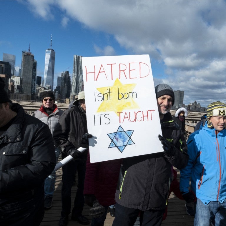 Photo of people marching on a bridge under a blue sky, man holding a poster sign that says 'hatred isn't born it's taught' with a Jewish star on the poster