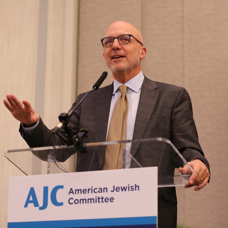 Image of AJC CEO Ted Deutch in suit and tie, speaking, wearing glasses, in front of a clear lectern