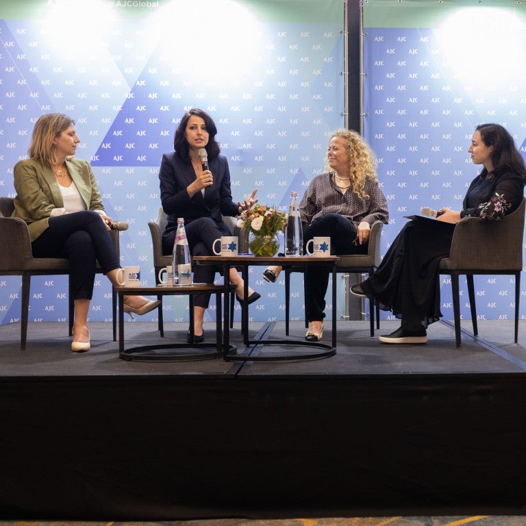 four people, Aviva Steinberger, Gadeer Kamal-Mreeh, Ayelet Nahmias-Verbin, Reva Gorelick, sitting on a black stage in front of a blue step and repeat background with the American Jewish Committee (AJC) logo, two tables with water bottles on top, and on the right side of the stage is a blue AJC-branded podium with a microphone
