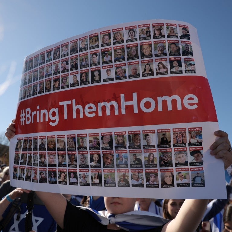 someone holding up a large sign that covers most of the frame. the sign is a mosaic of images of over 200 people kidnapped by hamas. there is a red banner going through the middle that reads in white text, #BringThemHome. the background is a blue sky.