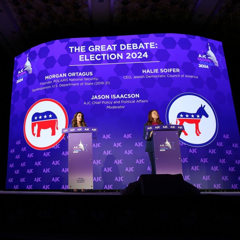 On a stage, there's a large desk and two podiums. A man, Jason Isaacson, is sat at the desk. Two women, Morgan Ortagus on the left and Halie Soifer on the right, stand at podiums on a stage with a backdrop that says "AJC Global Forum 2024" and "The Great Debate: Election 2020," followed by their names
