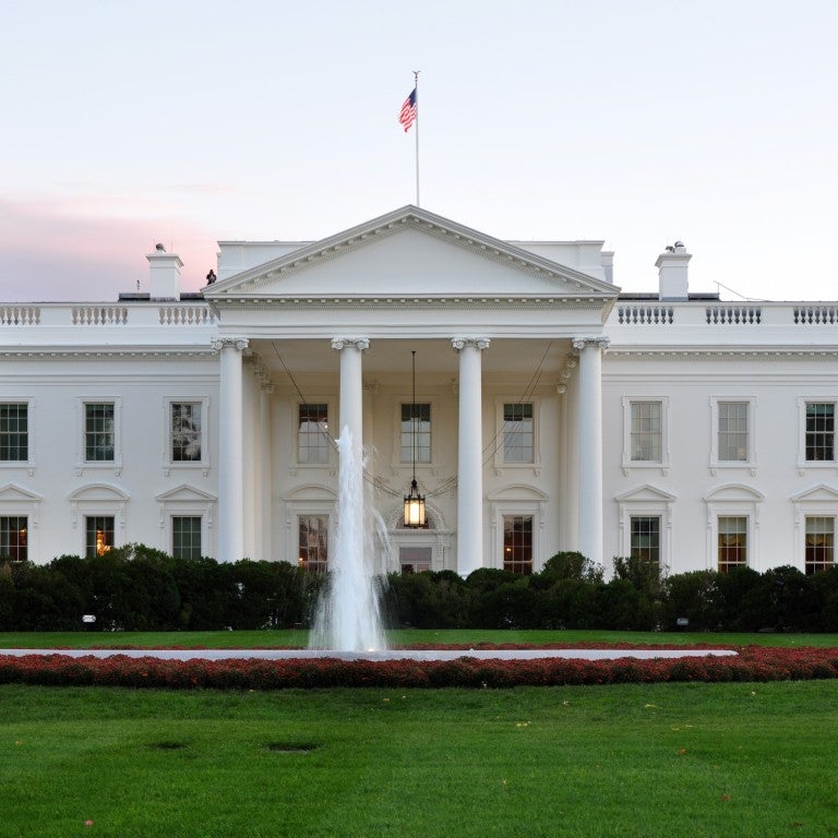 A scenic image of the White House with an American flag on the roof, featuring a fountain and green lawn.