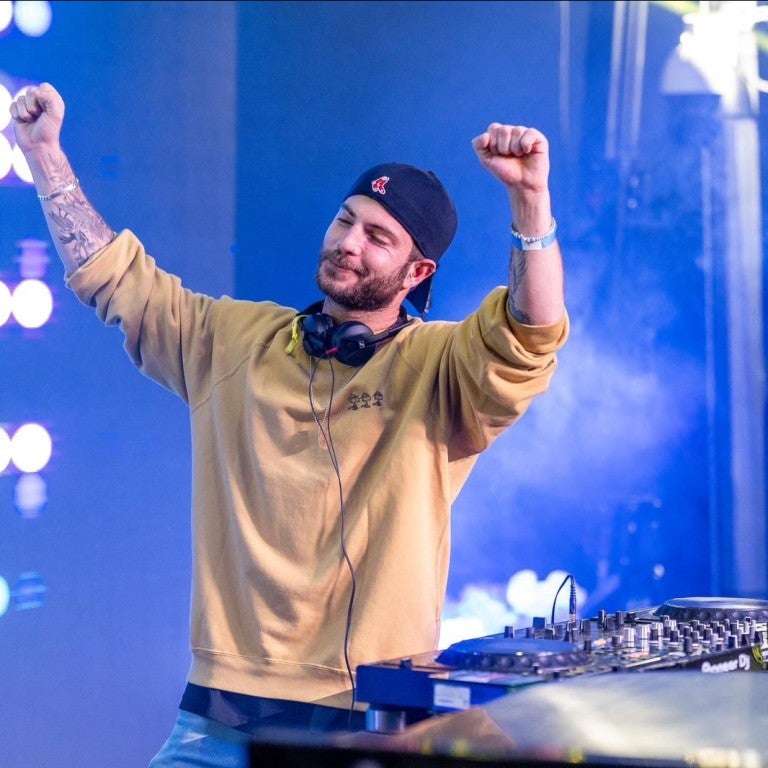 Israeli DJ Daniel Vaknin, in a yellow sweater stands with his arms raised, expressing joy or celebration. 