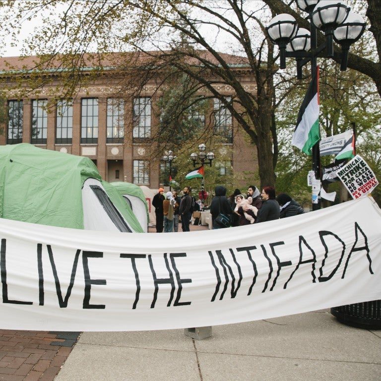 White banner reading "long live the intifada" in all caps, black lettering, in front of an encampment on University of Michigan's Ann Arbor, MI campus Diag.