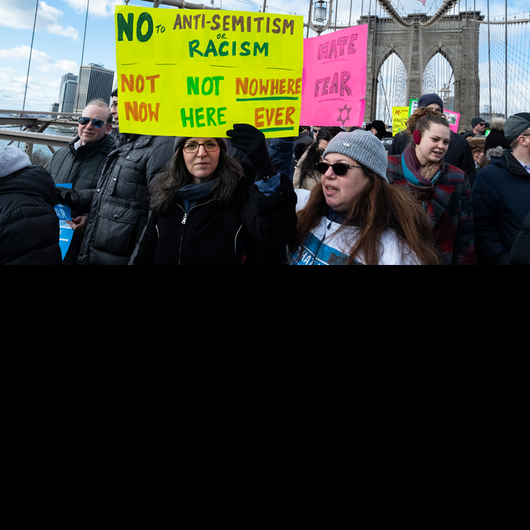 Woman marching across the Brooklyn Bridge with a sign saying "No to Antisemitism or Racism -- Not Now, Not Here, Nowhere Ever